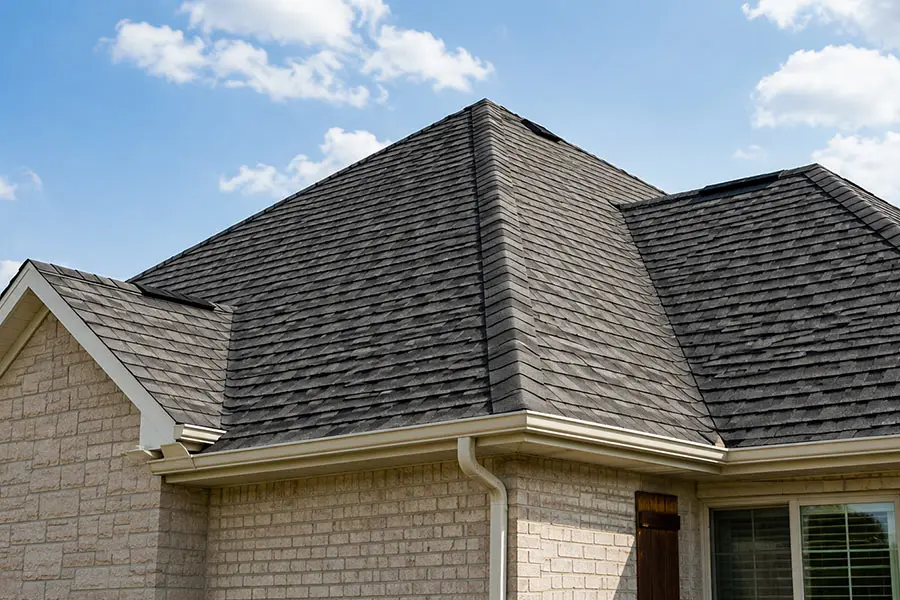 Residential home with a dark asphalt shingle roof, light brick exterior, gutters, and downspouts under a blue sky. Residential home with a dark asphalt shingle roof, light brick exterior, gutters, and downspouts under a blue sky.